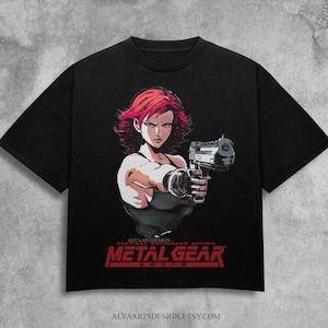 May include: Black t-shirt featuring a stylized anime illustration of a woman with red hair holding a gun. The shirt has the text "METAL GEAR SOLID" in red, with additional text above and below the logo. The Etsy shop URL is at the bottom.