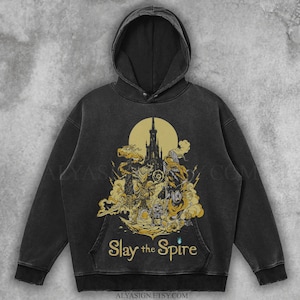 May include: Black hooded sweatshirt featuring a gold and yellow graphic of a castle, characters, and the text "Slay the Spire." The design is centered on the front, with a kangaroo pocket. The sweatshirt has a vintage, washed look.