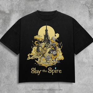 May include: Black t-shirt featuring a golden illustration of a castle, characters, and the text "Slay the Spire." The design includes a full moon and intricate details. The Etsy shop name is visible at the bottom.