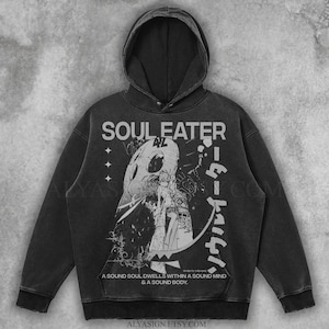 Può includere: Felpa con cappuccio nera con la scritta "SOUL EATER" e caratteri giapponesi. La grafica frontale include un'illustrazione dettagliata con figure e testo. La felpa ha un aspetto vintage e lavato, con cappuccio e coulisse.