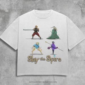 Pode incluir: Camiseta branca com um design de fantasia. A camiseta apresenta quatro personagens ilustrados e o texto "Slay the Spire" em dourado. Os personagens são diversos, incluindo um guerreiro e uma figura com túnica. O design é centralizado.