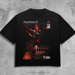 May include: Black t-shirt with a BloodRayne video game graphic. The design includes the PlayStation 2 logo, a red-haired character with weapons, and Japanese text. The shirt has a crew neck and short sleeves.