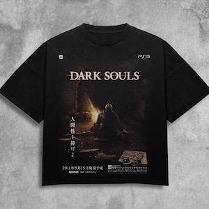 May include: Black t-shirt featuring the "DARK SOULS" logo and a graphic of a figure in a dark setting. The shirt includes Japanese text and a PS3 logo. The design is printed on a black, short-sleeved tee.