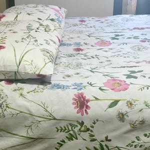 May include: White floral bed linens featuring a pillow and a fitted sheet. The pattern includes pink, blue, and white flowers with green stems and leaves. The bedding set is designed with a botanical theme.