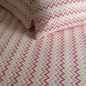 May include: Two pillows and a sheet with a repeating chevron pattern in red, peach, and white. The bedding has a zig-zag design, creating a visually appealing texture. The overall theme is home decor.