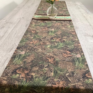 May include: A camouflage-patterned table runner with a design of leaves, pine needles, and pine cones. A vase of orange and yellow flowers sits on the runner. The runner has a brown and green striped border.