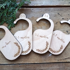 Peut inclure: Quatre cintres en bois avec des étiquettes pour nouveau-né, 0-3 mois, 3-6 mois et 6-9 mois. Chaque cintre est décoré d'un motif de feuilles.