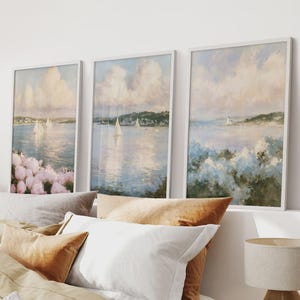 May include: Three framed wall art prints featuring coastal landscape paintings. Each print displays a calm body of water with sailboats, distant shorelines, and soft, cloudy skies. The foregrounds show clusters of pink and blue flowers.