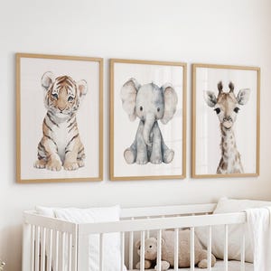 May include: Three framed watercolor prints featuring a tiger cub, an elephant, and a giraffe. The artwork has a soft, pastel color palette and is displayed above a white crib. The frames are a light wood color.