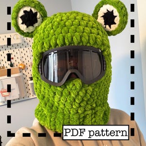 Könnte beinhalten: Eine leuchtend grüne, gehäkelte Sturmhaube mit großen, runden, ohrenartigen Akzenten. Die Sturmhaube hat eine schwarze Skibrille und den Text "PDF pattern" in einem weißen Feld. Der Hut ist aus weichem, strukturiertem Garn gefertigt.