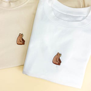 May include: Two crew neck sweatshirts, one cream and one white, each embroidered with a brown bear. The bear is sitting with its legs crossed. The sweatshirts are made of a soft material.