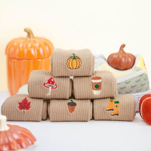 May include: A stack of six pairs of beige socks with embroidered autumn-themed designs, including a pumpkin, mushroom, acorn, coffee cup, and boots. The socks are arranged in a pyramid shape on a white surface.