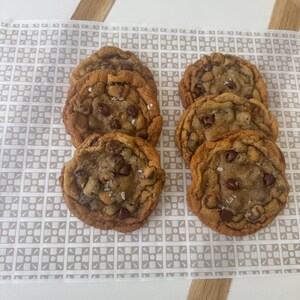 Puede incluir: Seis galletas de chocolate con chispas sobre una superficie con un patrón blanco y marrón. Las galletas son de color marrón dorado y tienen chispas de chocolate y sal marina en la parte superior.