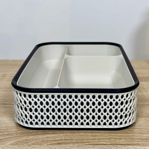 May include: A rectangular white and black organizer with three compartments. The exterior features a decorative openwork design. The rim is black, and the interior is white. A beige container is visible in the background.