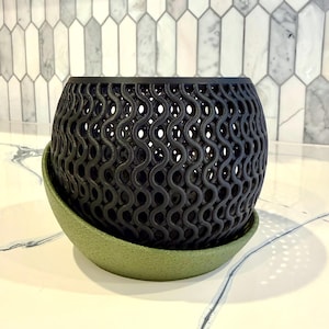 May include: A black, openwork planter with a wavy pattern sits on a green saucer. The planter has a cylindrical shape and is made of a dark material. The saucer is a solid, muted green color.