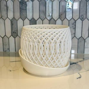 May include: A white, latticework-style planter with a matching saucer. The planter has a rounded shape with a woven design, and the saucer is a simple, curved dish. The background features a decorative tile backsplash.