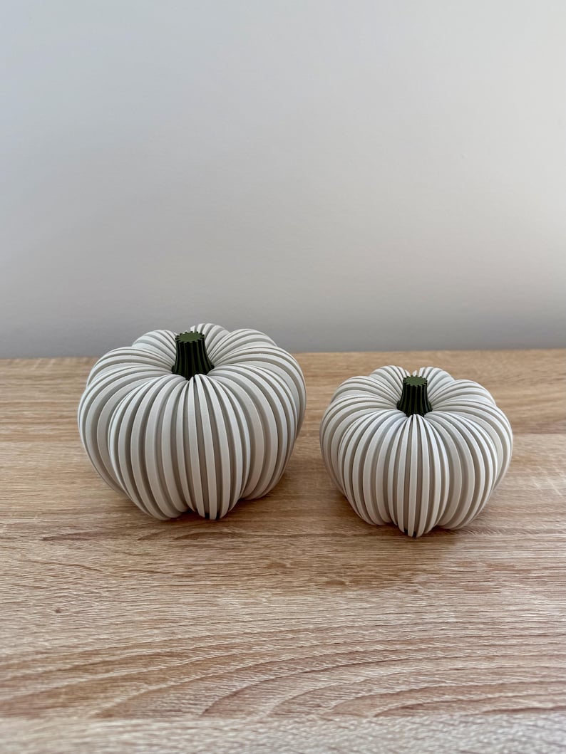 May include: Two decorative pumpkins with a ribbed design and green stems. The pumpkins are white with gray stripes and sit on a wooden surface. The pumpkins are different sizes, suggesting a set for fall decor.