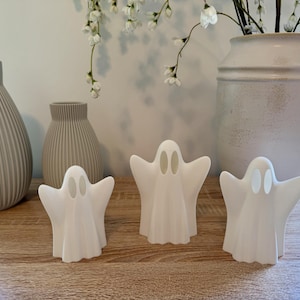 May include: Three white ghost figurines with oval eye cutouts are displayed on a wooden surface. Two gray vases and a large white vase with flowers are in the background. The ghosts have outstretched arms.