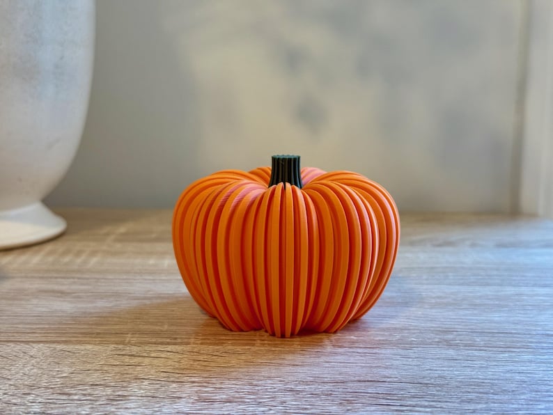 May include: An orange, ribbed pumpkin decoration with a black stem. The pumpkin is made of vertical slats, giving it a unique, modern look. Perfect for fall or Halloween decor.