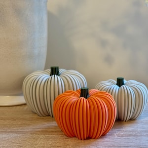 May include: Three decorative pumpkins in a still life arrangement. Two are white with green stems, and one is bright orange. The pumpkins are on a wooden surface, with a large white vase in the background.