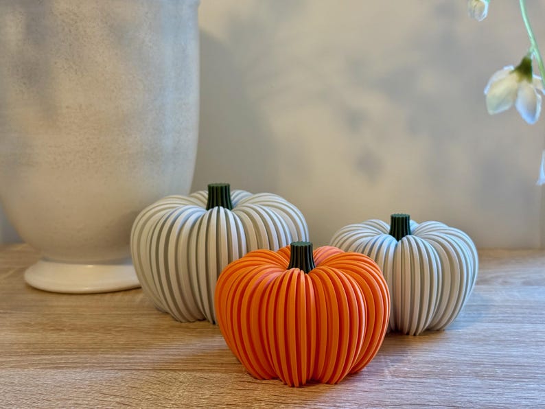 May include: Three decorative pumpkins in a still life arrangement. Two are white with green stems, and one is bright orange. The pumpkins are on a wooden surface, with a large white vase in the background.