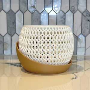 May include: A white, open-weave ceramic planter with a rounded shape, sitting on a brown, curved saucer. The planter has a decorative, lattice-like pattern. The background features a patterned tile wall.