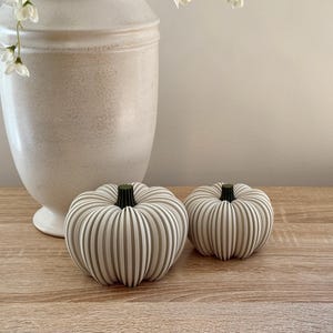 May include: Two decorative, cream-colored pumpkins with vertical grooves and dark green stems sit on a wooden surface. A large, off-white vase with a rounded shape is behind the pumpkins. White flowers are in the vase.