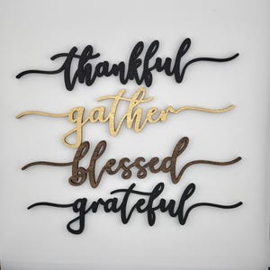May include: Four wooden wall art pieces with the words "thankful", "gather", "blessed", and "grateful" in a cursive font. The words are in black, gold, brown, and black, respectively. The pieces are designed for home decor.