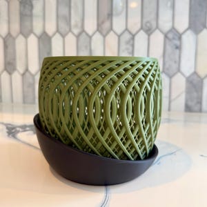May include: A green, lattice-patterned planter sits atop a dark, round saucer. The planter has a woven design, and the color is a muted green. The saucer is a dark, matte color. The background is a light-colored surface with a patterned wall.