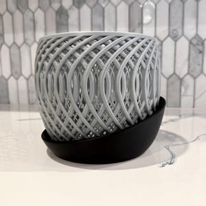 May include: A gray, woven-patterned planter pot with a black saucer. The pot has a unique, open design, allowing for air circulation. The planter is designed to hold small plants and is suitable for indoor or outdoor use.