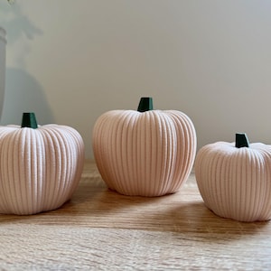 May include: Three decorative pumpkins in varying sizes. The pumpkins are a soft pink color with vertical ribbed detailing. Each pumpkin has a small, dark green stem. They are arranged on a light-colored wooden surface.