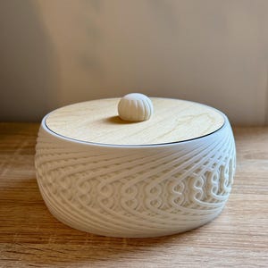 May include: A white decorative container with a wooden lid. The container has a detailed, woven pattern around the sides. The lid has a small, round knob. The container is sitting on a wooden surface.