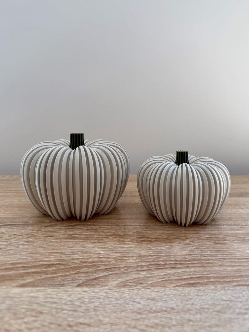 May include: Two decorative pumpkins with a ribbed design. The pumpkins are light gray with black stems and sit on a wooden surface. One pumpkin is larger than the other, creating a fall-themed display.