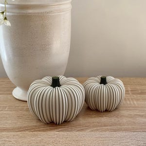 May include: Two decorative pumpkins with ribbed detailing and dark green stems. The pumpkins are off-white and sit on a wooden surface. A large, off-white vase is in the background. The pumpkins are a fall or autumn themed decoration.