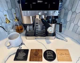 May include: A stainless steel Ninja espresso machine with a black control panel sits on a countertop. Clear glass bottles with gold tops and a white mug are nearby. Four coasters with coffee-themed designs are in front.