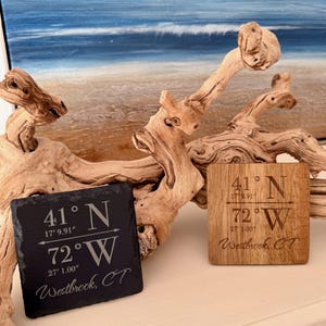 May include: Two square coasters, one black slate and one wood, each engraved with coordinates and "Westbrook, CT." The black coaster displays "41° N 17'9.91" 72° W 27'1.00". The wooden coaster has the same text. Driftwood and a beach painting are in the background.