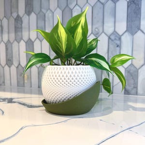 May include: A white, spherical, 3D-printed planter with a woven design, holding a green and yellow plant. The planter sits on a green, leaf-shaped saucer. The background features a gray and white tiled wall.