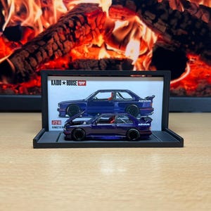 Może przedstawiać: Miniaturowy fioletowy model samochodu z czarnymi kołami i napisem "ADVAN" jest prezentowany w czarnej ramie. Rama zawiera również niebieską ilustrację samochodu i napis "KAIDO * HOUSE". W tle widać kominek z płonącymi kłodami.