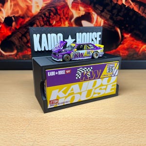 Może przedstawiać: Miniaturowy fioletowo-żółto-biały model samochodu Kaido House BMW E30 M3 KW V1 z numerem 95 z boku. Samochód stoi na czarnym stojaku wystawowym z logo Kaido House. W tle widać kominek.