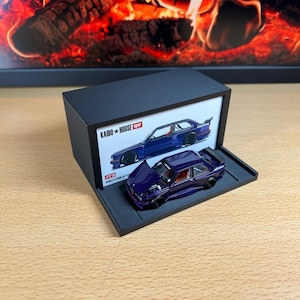 May include: A diecast model car in a black display case. The car is a deep purple with black wheels and red interior details. The display case has the text "KAIDO + HOUSE" and an image of a blue car.