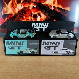 Może przedstawiać: Dwa miniaturowe modele samochodów, jeden turkusowy i jeden srebrny, prezentowane na czarnych pudełkach z logo "MINI GT". Turkusowy samochód ma kolorowy design, a srebrny jest elegancki. Pudełka zawierają zdjęcia samochodów.