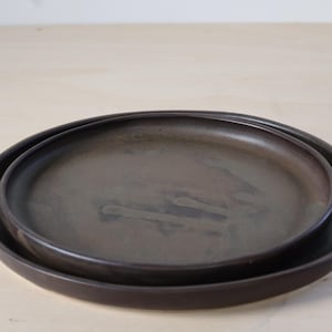 May include: Two dark brown ceramic plates, one nested inside the other. The plates have a simple, modern design with a slightly textured surface. The larger plate is approximately 25 cm in diameter.