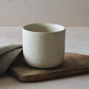 Puede incluir: Una taza cilíndrica de cerámica con un acabado moteado en color blanco roto. La taza tiene un borde redondeado y liso con paredes rectas. Se sitúa junto a un paño de lino gris plegado y una tabla de cortar de madera con mango.