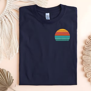 Könnte beinhalten: Gefaltetes marineblaues T-Shirt mit einem Retro-Sonnenuntergangsdesign auf der linken Brust. Das Sonnenuntergangs-Design hat horizontale Streifen in Orange, Gelb, Türkis und Rot.