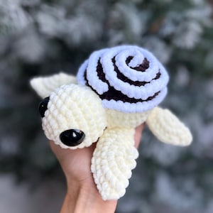 Puede incluir: Una tortuga de crochet hecha a mano. La tortuga tiene un cuerpo color crema, ojos negros y un caparazón con un diseño en espiral marrón y blanco. El juguete suave y afelpado es perfecto para niños.