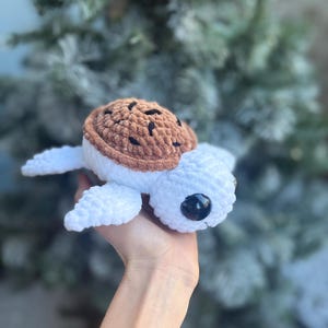 May include: A handmade crochet turtle with a white body, brown shell, and black eyes. The turtle is held in a hand against a blurred background of green and white. The turtle's shell has dark brown spots.