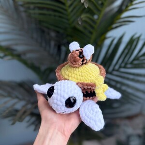 May include: A handmade crochet turtle with a bee on its back. The turtle is white with black eyes and white flippers. The bee is brown and yellow, perched on a yellow honeycomb. The turtle is being held in a hand.