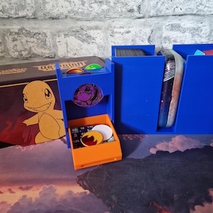 May include: A blue and orange card game organiser with a box featuring a cartoon character. The organiser has compartments for cards, coins, and other game accessories. The box has the text "Pokemon" and a cartoon character.
