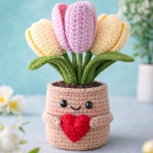 May include: A crocheted flower pot with a smiling face and a red heart. The pot holds three crocheted tulips in shades of pink and yellow, with green leaves. The pot is tan and has small arms.