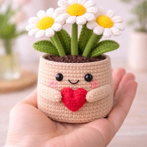 May include: A crocheted flower pot with three white daisies with yellow centers and green stems. The pot is beige with a smiling face, pink cheeks, and a red heart. The pot has small arms and is held in a hand.